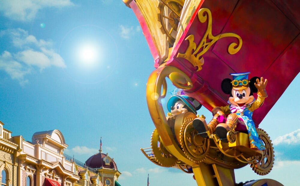 Topolino e Minnie in costumi colorati cavalcano uno stravagante carro da parata di ispirazione steampunk sotto un cielo azzurro e soleggiato a Disneyland Paris, con gli edifici di Main Street sullo sfondo.