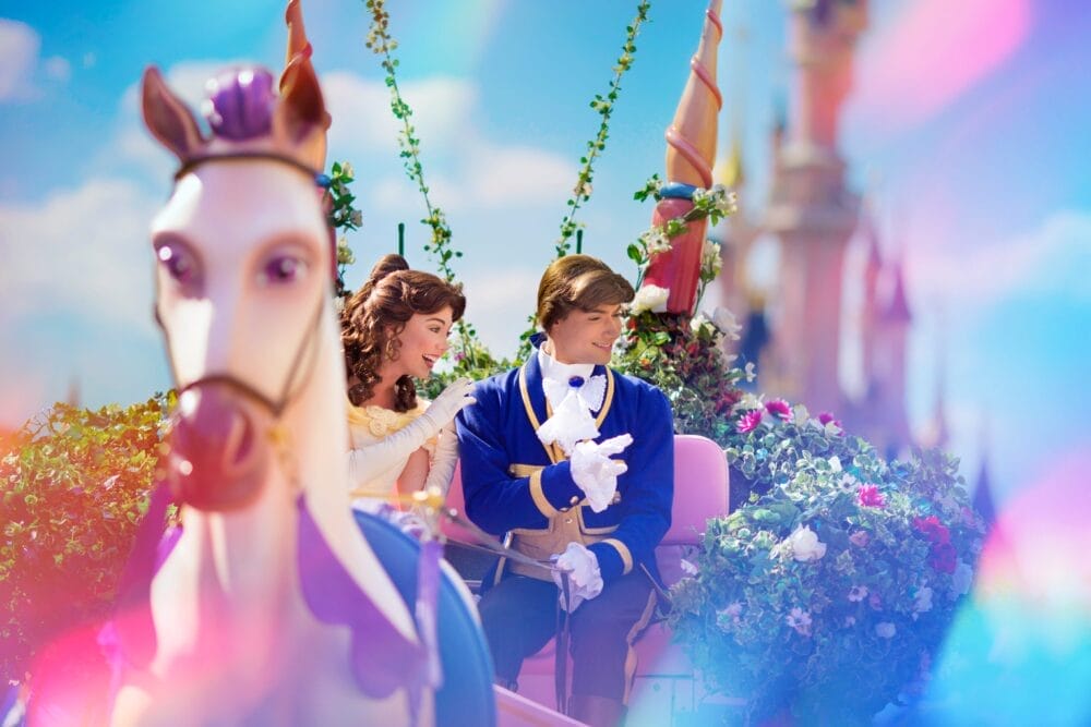 Una donna nei panni di Belle e un uomo nei panni della Bestia (in forma umana) cavalcano una carrozza adornata di fiori con un cavallo bianco a Disneyland Paris, passando davanti a luci luminose e a un castello, catturando la magia subito dopo Ingresso al Parco.