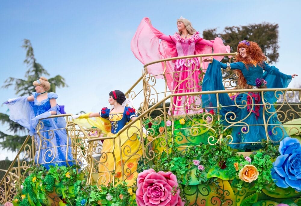 Quattro donne vestite da principesse in abiti colorati si trovano su un carro da parata decorato a Disneyland Paris, circondato da ringhiere dorate e fiori vivaci, e salutano la folla sotto un cielo luminoso vicino all'Ingresso al Parco.