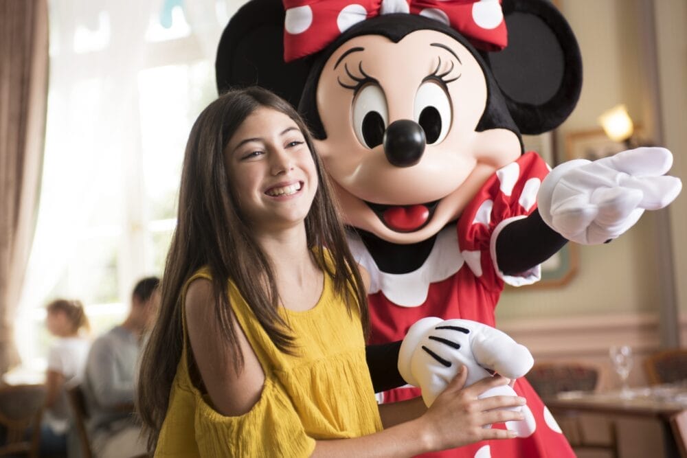 Una ragazza sorridente con un vestito giallo sta accanto a Minnie Mouse, che indossa un fiocco e un vestito a pois rossi, catturando la gioia di un Soggiorno a Disneyland Paris in un ambiente interno luminoso e allegro.