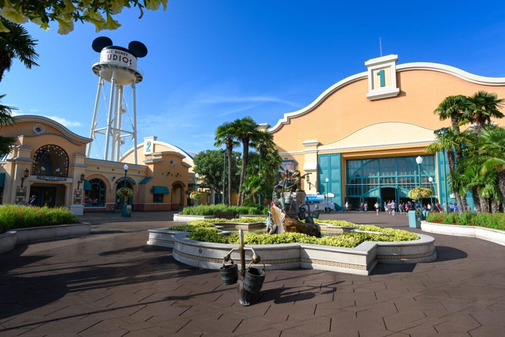 Giornata di sole all'Ingresso al Parco di Disneyland Paris, con edifici gialli, una fontana con la statua di un personaggio, palme e un'alta torre d'acqua con le orecchie di Topolino con la scritta "STUDIO".