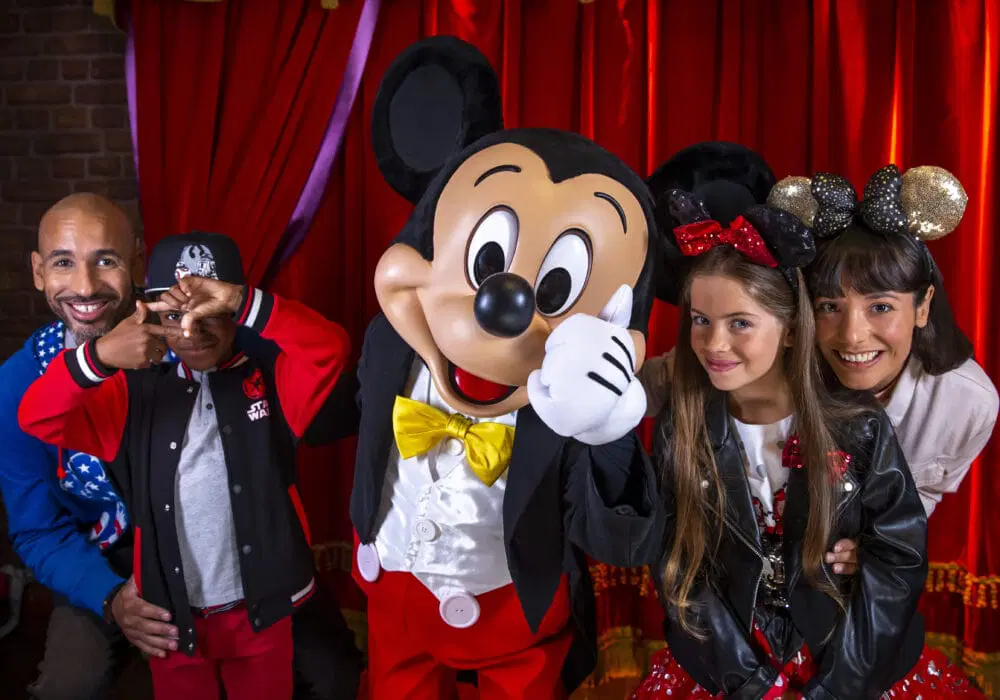 Una famiglia di quattro persone posa felice con Topolino davanti alle tende rosse di Disneyland Paris. Topolino saluta con il suo costume classico, mentre la famiglia sorridente - alcuni con le orecchie di Topolino e i fiocchi di Minnie - festeggia il suo ingresso al parco.