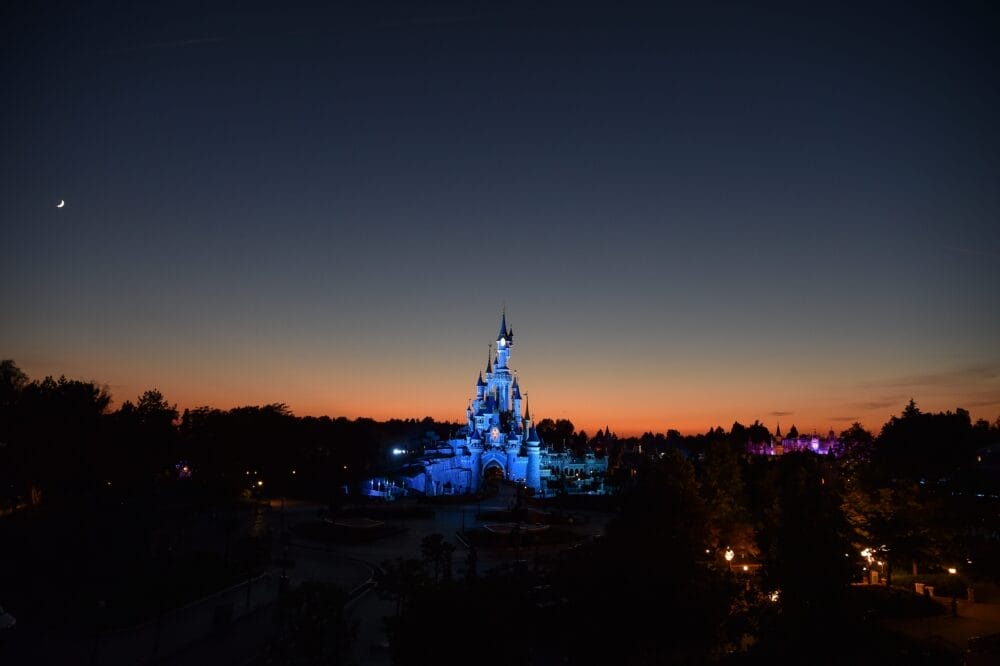 Il castello delle fiabe di Disneyland Paris, illuminato a giorno, si staglia contro una sagoma scura di alberi e un cielo colorato al tramonto, con una luna crescente visibile nell'angolo in alto a sinistra.