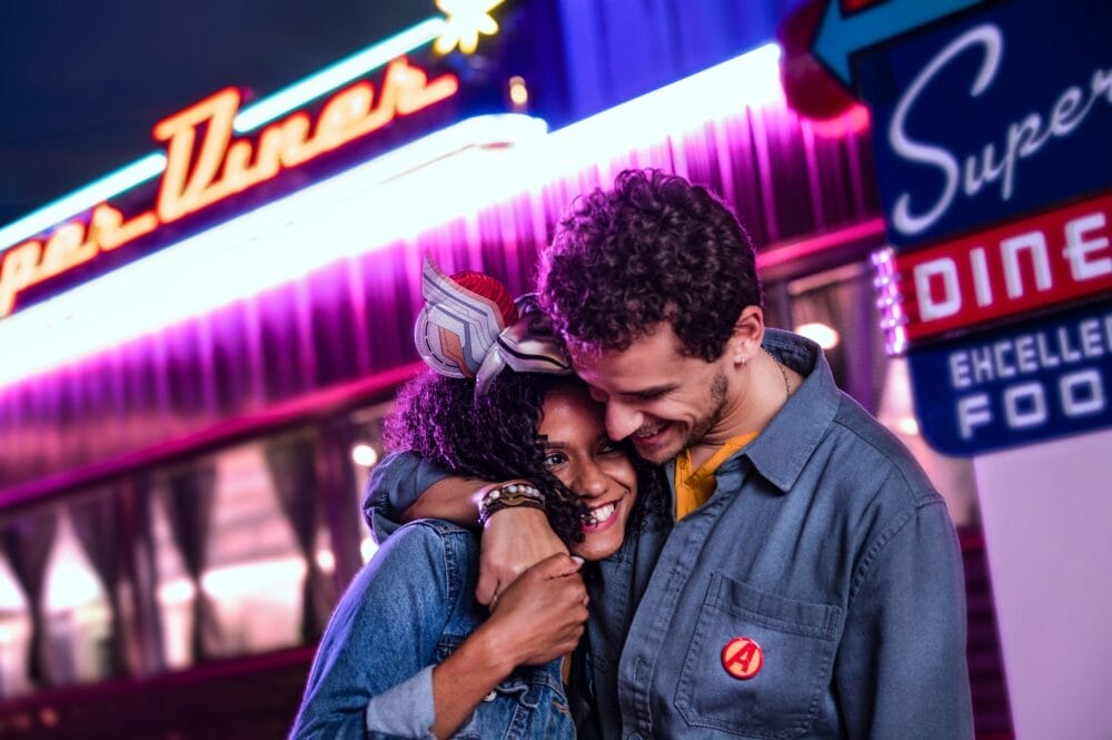 Una coppia sorridente si abbraccia fuori da un locale retrò illuminato di notte, con luci colorate e insegne al neon che brillano come quelle di Disneyland Paris, catturando la magia di un Soggiorno indimenticabile.