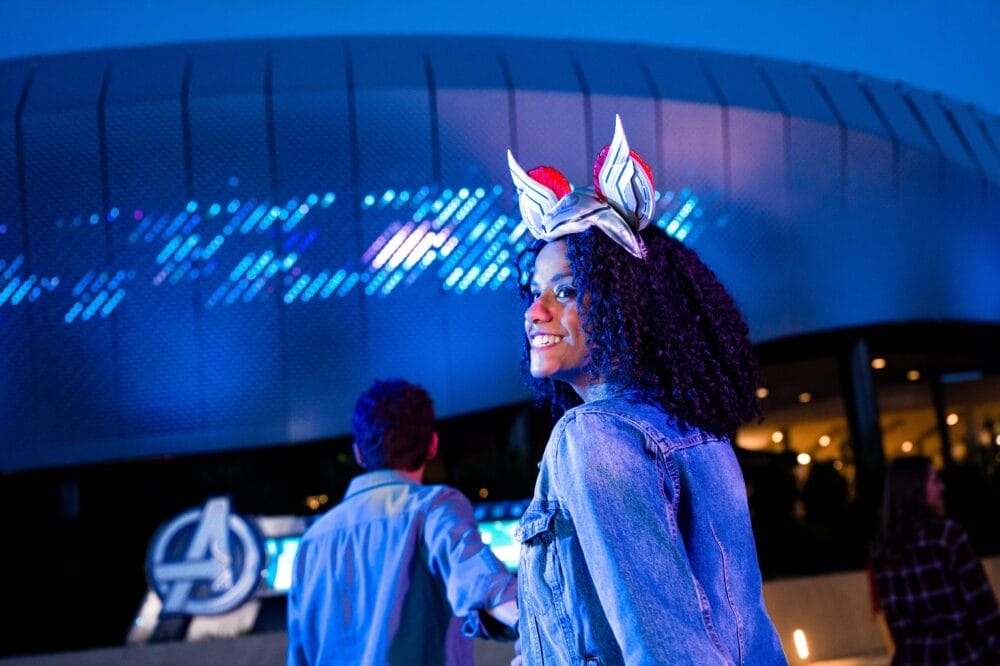 Una donna sorridente con una giacca di jeans e una fascia per capelli a tema Avengers si trova all'esterno dell'ingresso al parco illuminato di notte, tenendo per mano qualcuno. Il logo degli Avengers brilla sullo sfondo, catturando la magia di Disneyland Paris.