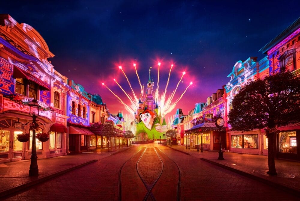 Una strada illuminata conduce al castello colorato di Disneyland Paris di notte, con i fuochi d'artificio che scoppiano nel cielo. Gli edifici brillano di luci vivaci e la strada vuota crea un'atmosfera magica per il vostro Soggiorno.