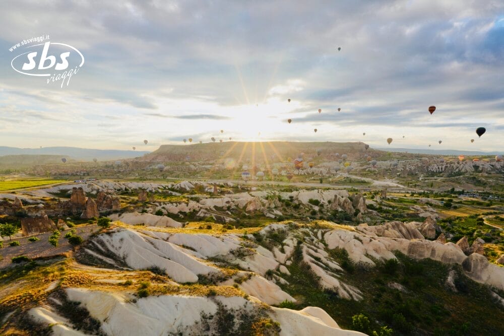 Numerose mongolfiere fluttuano sopra un paesaggio roccioso e verde all'alba in questa bozza automatica, con la luce del sole che filtra attraverso le nuvole. Il terreno presenta formazioni rocciose uniche e verde sparso.