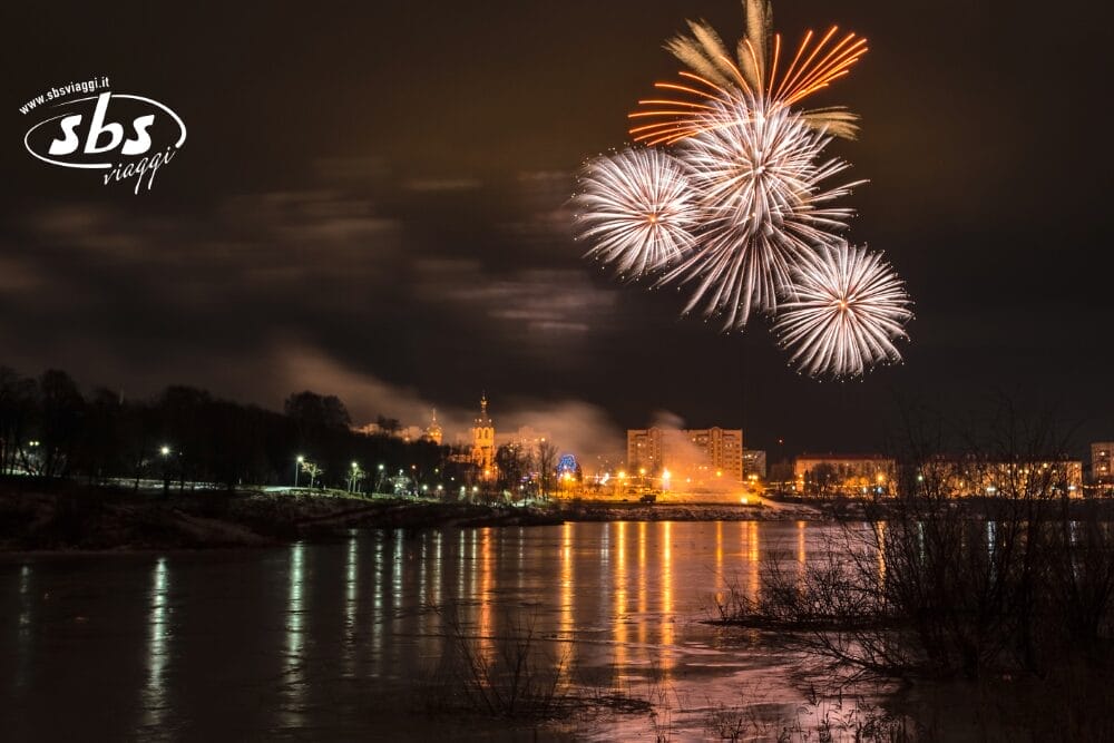 I fuochi d'artificio esplodono nel cielo notturno su un paesaggio urbano che si riflette su un fiume tranquillo. Gli edifici e le luci risplendono di luce calda, con alberi senza foglie lungo il bordo dell'acqua. Il logo "sbs viaggi" e il testo Bozza automatica appaiono nell'angolo.