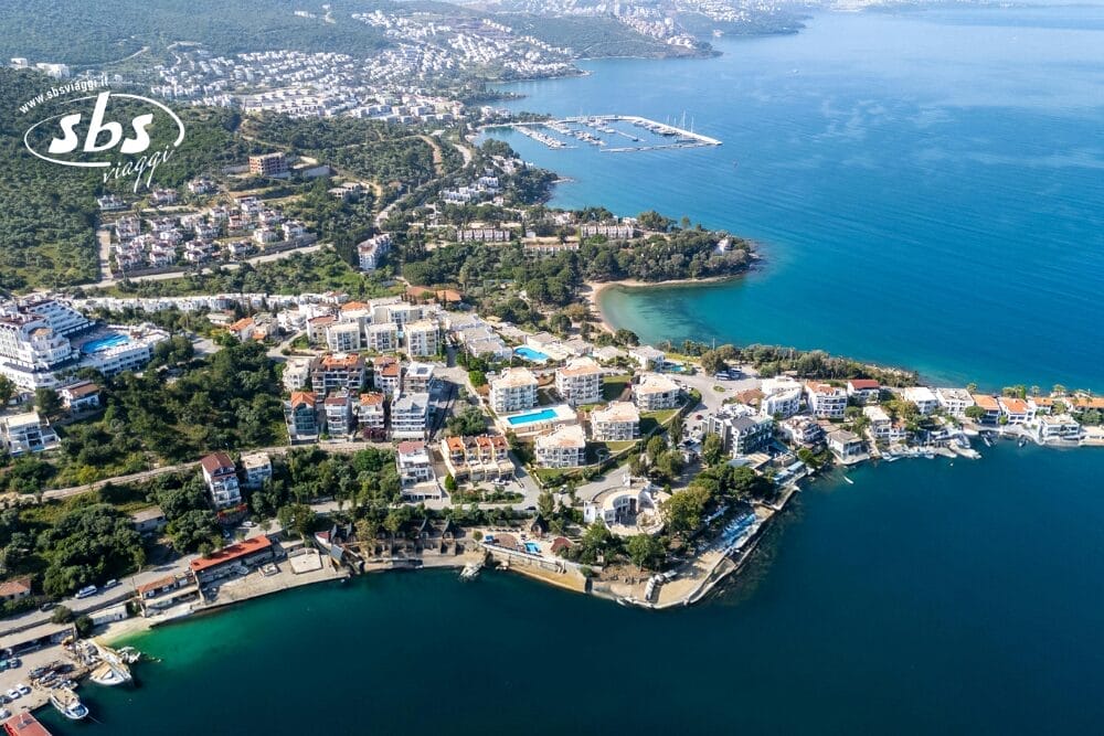 Veduta aerea di una città costiera con edifici bianchi, alberi verdi, piccole spiagge e un porticciolo con barche su acque azzurre e trasparenti: questa scena pittoresca offre una vera e propria bozza automatica in mezzo a un paesaggio collinare.