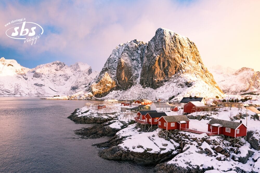 Cabine rosse coperte di neve si trovano su una riva rocciosa accanto ad acque tranquille, con una montagna imponente sullo sfondo e Bozza automatica che si perde nel cielo parzialmente nuvoloso in un paesaggio invernale.