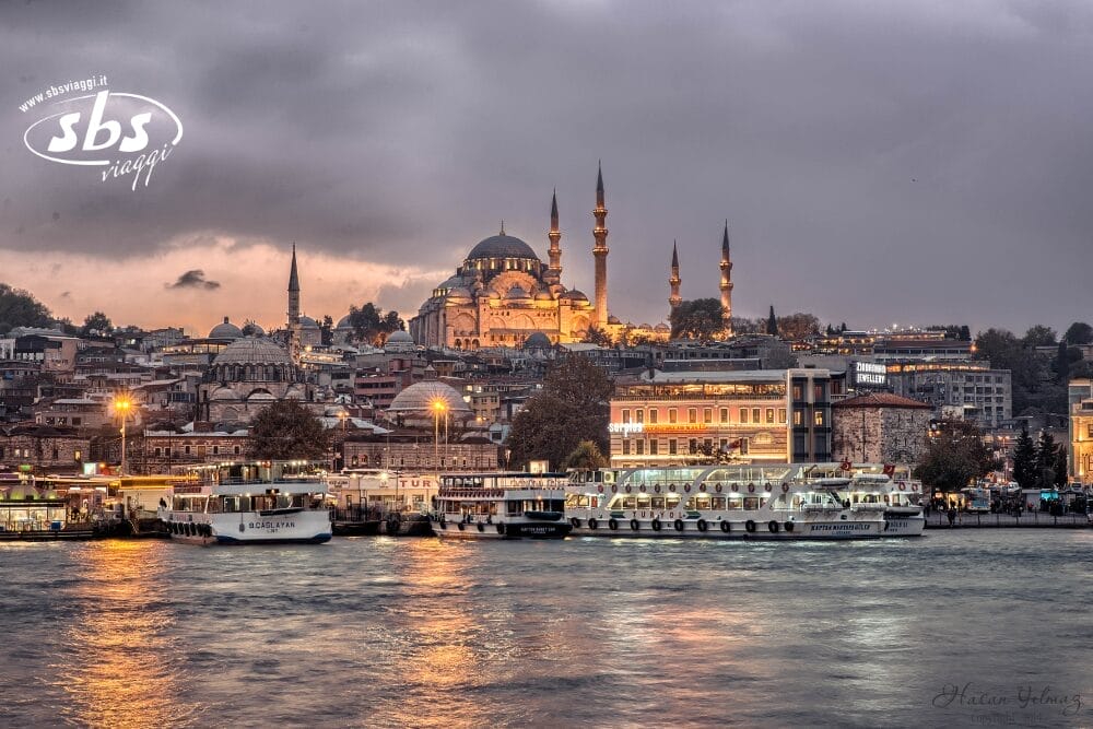 Una veduta del lungomare di Istanbul al tramonto, con le moschee illuminate, gli edifici della città e le barche attraccate lungo la riva, che si riflettono sull'acqua sotto un cielo nuvoloso: una scena che ricorda una bozza automatica dalle tinte vivaci.