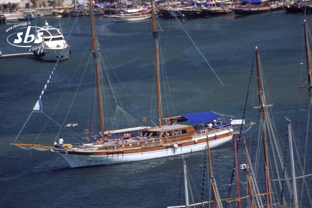 Un grande yacht a vela in legno con due alberi e un tettuccio blu naviga in un porto turistico, con la sua Bozza automatica che guida il percorso, mentre le persone si rilassano sul ponte. Sullo sfondo sono visibili altre imbarcazioni e il porto.