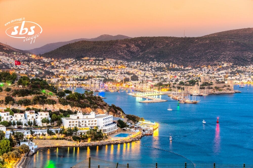 Una città costiera con edifici bianchi, un porto turistico pieno di barche e colline sullo sfondo al tramonto. La Bozza automatica delle luci del lungomare brilla sul mare azzurro e calmo, riflettendo la vivace energia della città.