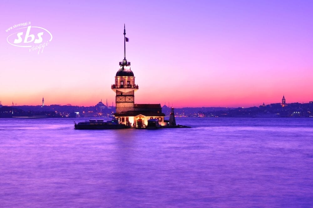 Una piccola torre illuminata si erge su un isolotto in acque calme al tramonto, incorniciata da un cielo vibrante di viola e rosa, dallo skyline della città sullo sfondo e da una scena tranquilla che ricorda un dipinto automatico di Bozza.