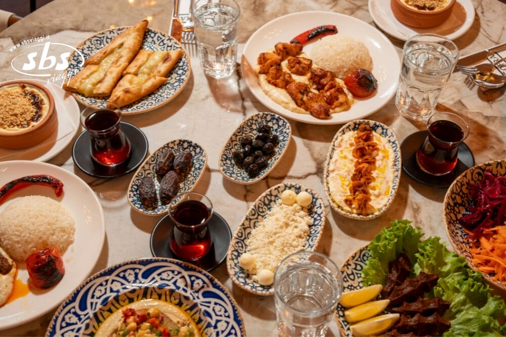 Tavola imbandita con vari piatti turchi, tra cui riso, spiedini di carne alla griglia, foglie d'uva ripiene di Bozza automatica, olive, datteri, insalata, baklava, pane e bicchieri di tè e acqua su piatti decorativi.