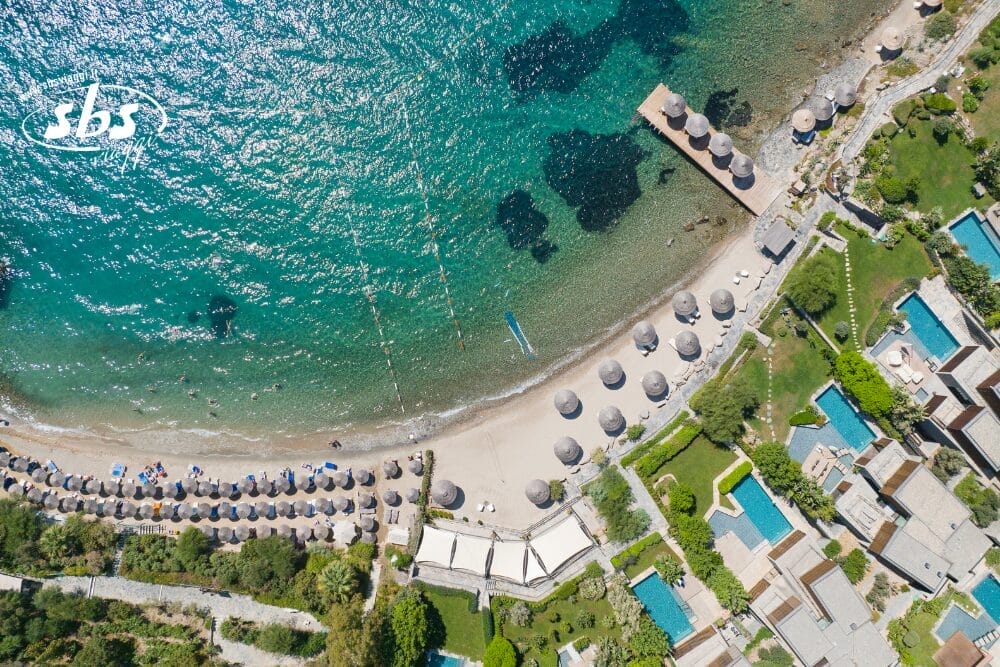 Veduta aerea di un resort costiero con acqua turchese, spiaggia sabbiosa fiancheggiata da ombrelloni, cabine e piscine circondate da verde ed edifici. La scena evoca una bozza automatica di relax mentre i nuotatori scivolano sulle rocce sottomarine visibili.