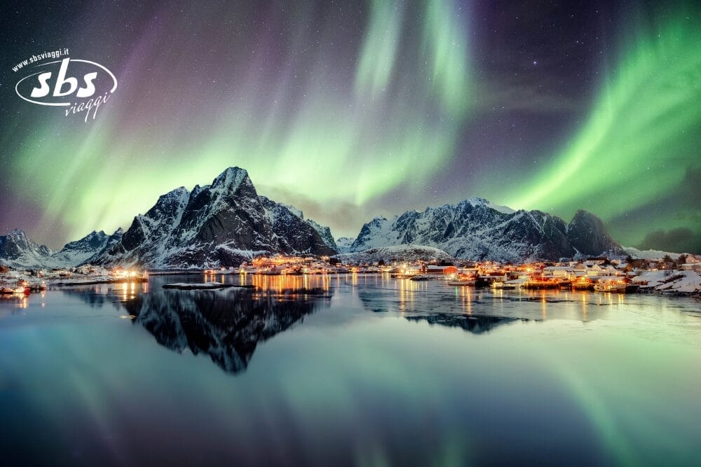 L'aurora boreale illumina il cielo notturno sopra le montagne innevate e un villaggio costiero norvegese, con riflessi nell'acqua calma. Le piccole case risplendono di luci calde. Il logo "sbs viaggi" e "Bozza automatica" appaiono nell'angolo in alto a sinistra.