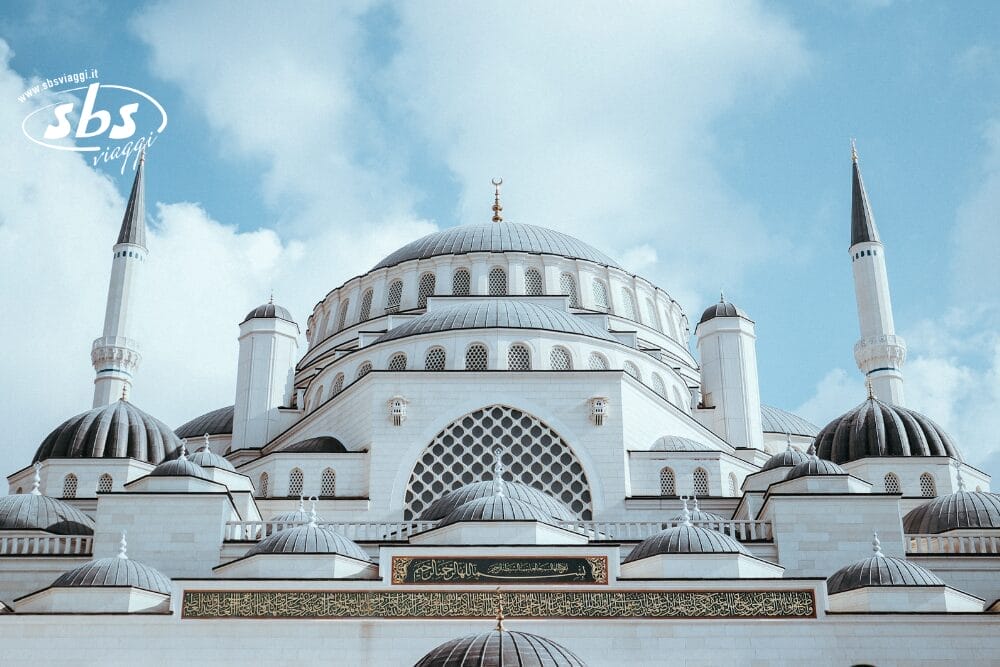 Una grande moschea bianca con cupole multiple e alti minareti si staglia contro un cielo azzurro parzialmente nuvoloso. Motivi automatici di bozza e calligrafia araba decorano l'edificio sopra l'ingresso.