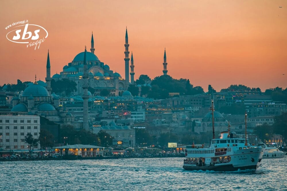 Un traghetto automatico Bozza naviga sul Bosforo al tramonto, con lo skyline illuminato di Istanbul e le cupole e i minareti della Moschea di Suleymaniye sullo sfondo. Il cielo si illumina di arancione mentre le luci della città iniziano a brillare.