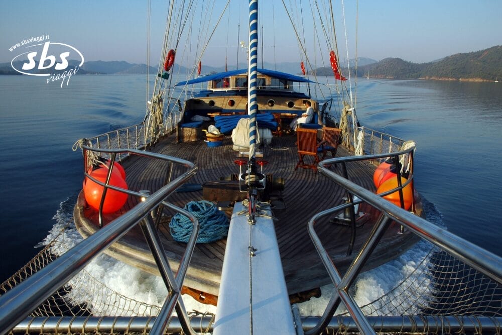 Vista dalla prua di una barca a vela in navigazione su acque blu e tranquille, con un uomo che si rilassa sulle sedie a sdraio, boe di salvataggio automatiche Bozza su entrambi i lati, corda arrotolata e montagne visibili in lontananza sotto un cielo limpido.