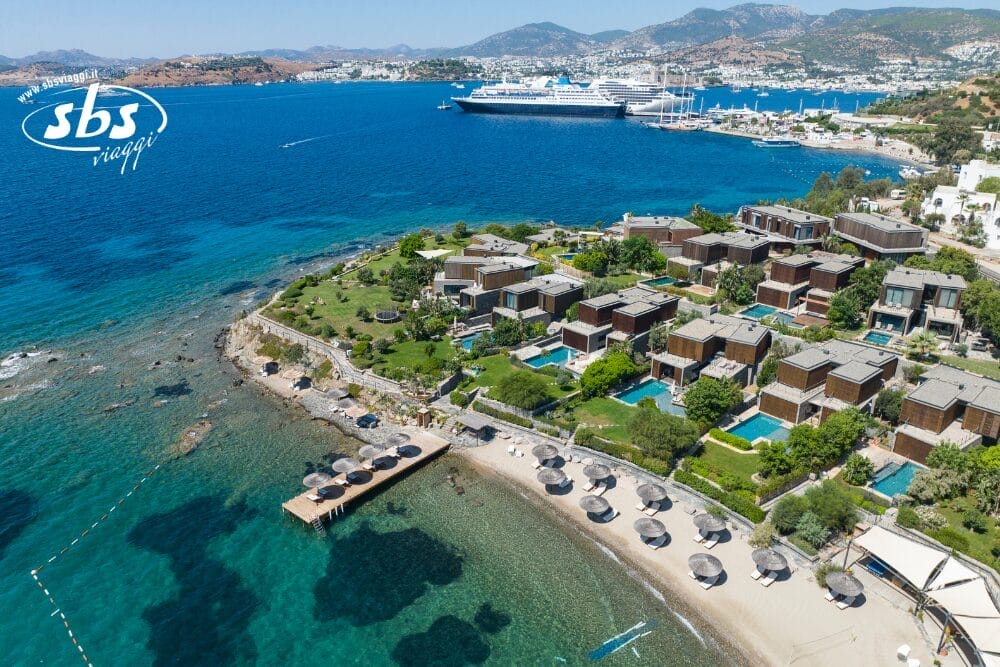 Vista aerea di un resort costiero con ville moderne, piscine private e una spiaggia sabbiosa con lettini. Un molo di legno si estende sul mare azzurro e limpido. Bozza automatica: nave da crociera e porto turistico visibili sullo sfondo accanto a colline lontane.