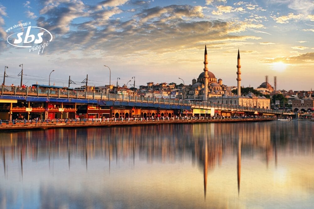 Una vivace scena sul lungomare al tramonto con la Nuova Moschea di Istanbul, con le sue cupole e i suoi minareti che si riflettono nell'acqua calma, e un ponte affollato di negozi e persone cattura lo spirito unico di bozza automatica della città.