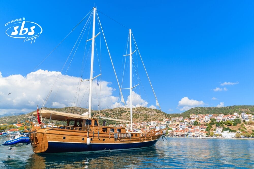 Una grande barca a vela in legno è ancorata in acque tranquille vicino a un villaggio costiero con case dai tetti bianchi e rossi, sullo sfondo di colline verdi e di un cielo blu brillante. Il logo "sbs viaggi" appare nell'angolo in alto a sinistra come parte di una Bozza automatica.