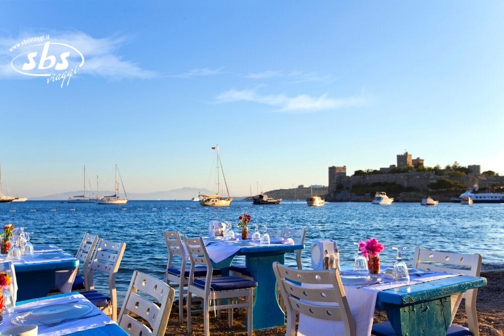 Tavoli da ristorante all'aperto con sedie bianche e blu disposti sul lungomare, affacciati su un mare calmo con barche a vela e un lontano castello di pietra sotto un cielo azzurro e limpido. I fiori decorano i tavoli, creando una cornice perfetta per un momento di Bozza automatica.