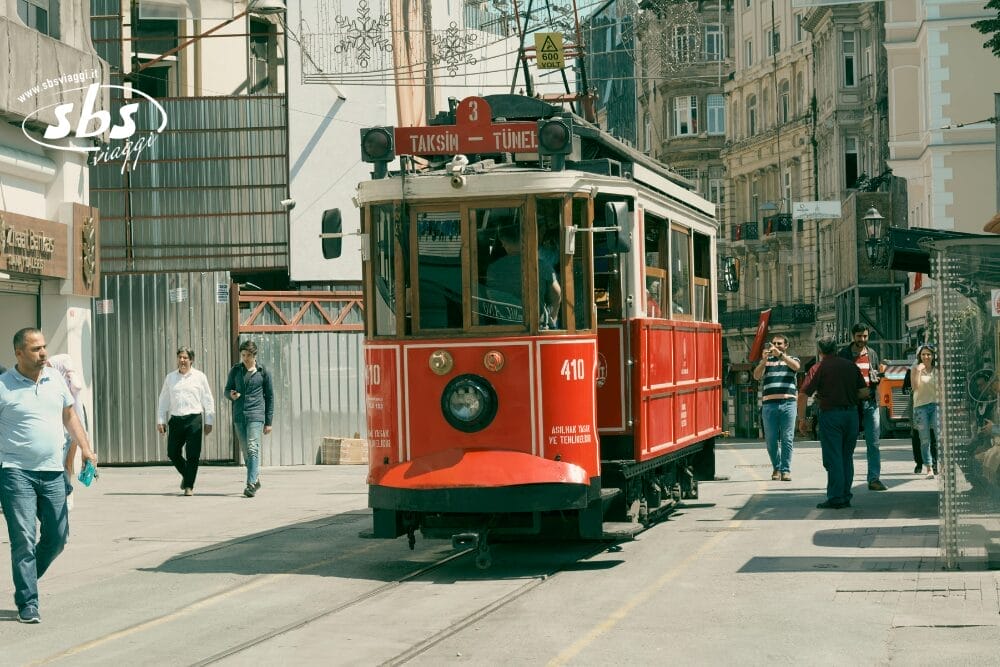 Un tram rosso d'epoca con la scritta "Taksim - Tünel" scivola lungo una strada cittadina fiancheggiata da edifici, mentre la gente passeggia nelle vicinanze in una giornata di sole a Istanbul, catturando il fascino della bozza automatica in mezzo alla vita urbana.
