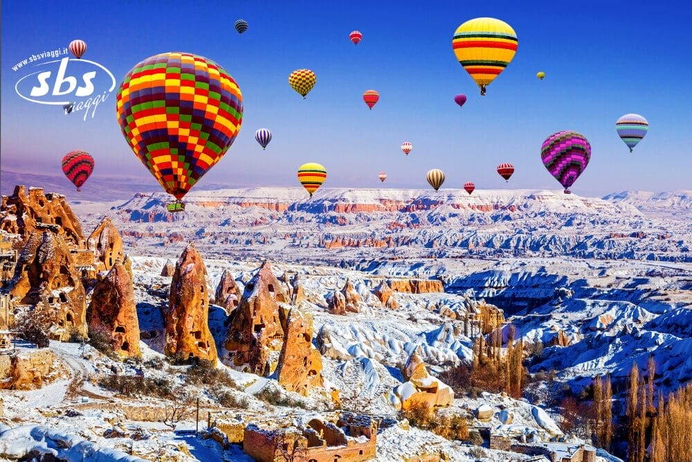 Mongolfiere di vari colori fluttuano sopra formazioni rocciose innevate in Cappadocia, Turchia, sotto un cielo blu brillante. Questa vista mozzafiato, come un capolavoro di Bozza, mette in risalto le uniche rocce appuntite e il paesaggio invernale.