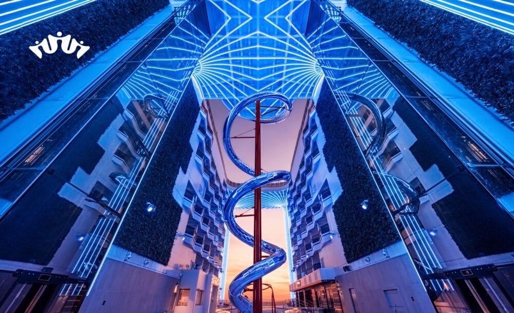 Un edificio futuristico con pareti a specchio, giardini verticali e un grande scivolo metallico a spirale al centro, illuminato da luci LED geometriche blu vibranti, che ricorda gli spazi innovativi a bordo di MSC World Asia in partenza da Civitavecchia.