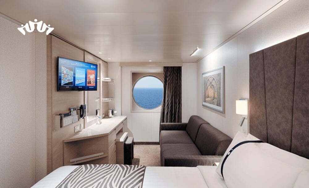 Moderna cabina di una nave da crociera MSC World Asia con letto, divano, TV a parete, scrivania e finestra rotonda con vista sulla crociera mediterranea. La camera è caratterizzata da colori neutri, arredamento minimal e opere d'arte incorniciate alla parete.