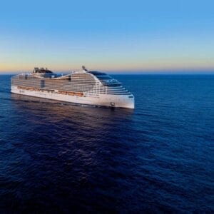 Una grande nave da crociera bianca MSC World Asia naviga sulle calme acque blu dell'oceano sotto un cielo limpido al tramonto, promuovendo un'indimenticabile crociera mediterranea. Un piccolo logo appare nell'angolo in alto a sinistra dell'immagine.