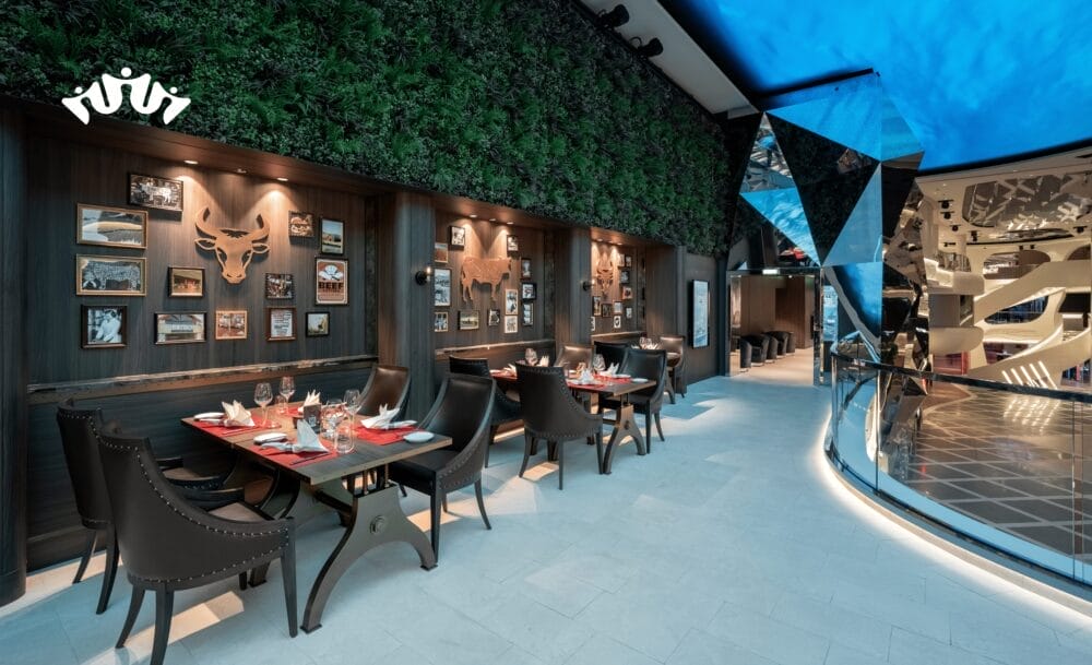 Un ristorante moderno con eleganti sedie e tavoli scuri, una parete di piante verdi, foto incorniciate e una struttura geometrica a specchio sotto un soffitto blu brillante: l'ideale per gli ospiti in viaggio verso la crociera mediterranea da Civitavecchia con MSC World Asia.