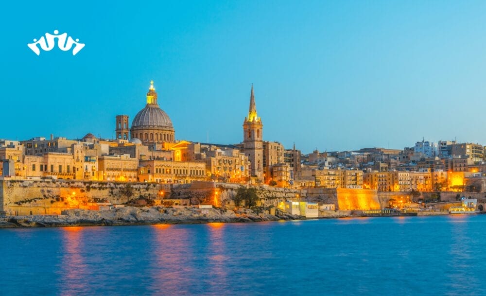 Un paesaggio urbano sul lungomare di Civitavecchia al tramonto, con edifici storici che risplendono, una cattedrale a cupola e un'alta guglia che si riflette sull'acqua blu e calma; il logo di una corona bianca galleggia sopra: uno scenario perfetto per una crociera Mediterraneo di MSC World Asia.