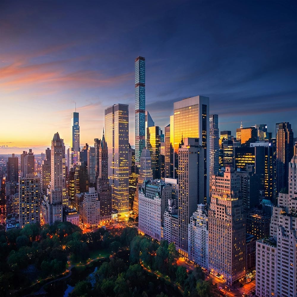 Lo skyline di una città al tramonto con alti grattacieli illuminati e Central Park in primo piano; il cielo è un gradiente di blu e arancione e le luci della città iniziano a brillare: una scena iconica degli Stati Uniti.