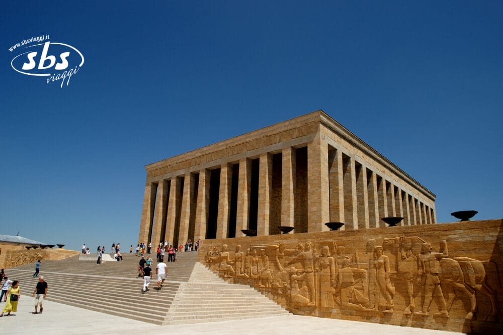 La gente sale ampi gradini verso l'Anıtkabir, il mausoleo di Mustafa Kemal Atatürk ad Ankara, in Turchia, con alte colonne e dettagliate sculture in pietra visibili sotto un cielo azzurro e limpido.