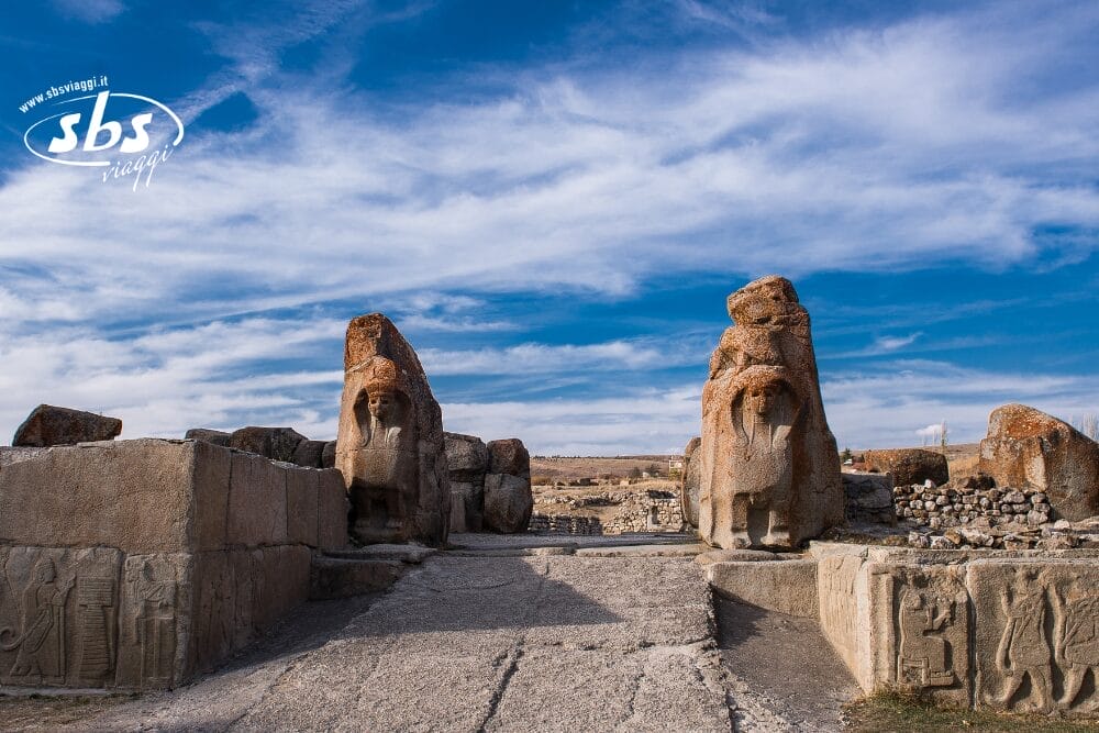Antica porta in pietra con due grandi statue di leoni e rilievi scolpiti sotto un cielo azzurro parzialmente nuvoloso, incastonata nel paesaggio aperto della Turchia - scopritela con SBSViaggi Stile e Turkish Airlines.