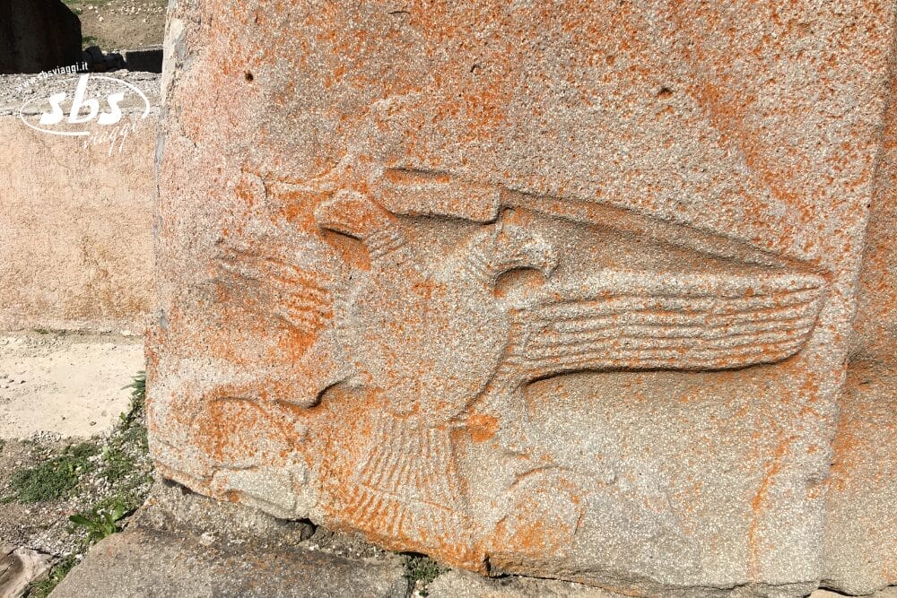 Antico rilievo in pietra raffigurante una figura alata con il volto di un uccello e le ali spiegate, scolpito in una pietra arancione e consumata dagli agenti atmosferici in Turchia. Alla base sono visibili erba e pietre.