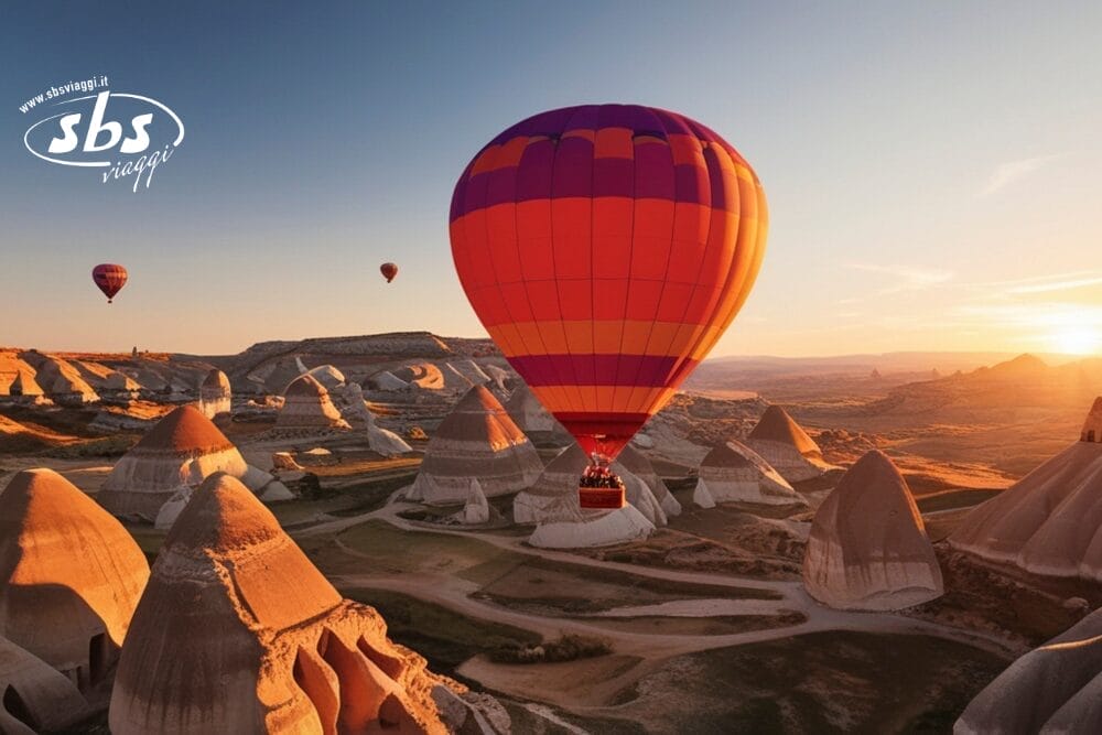 Una vivace mongolfiera fluttua sopra le singolari formazioni rocciose della Cappadocia, in Turchia, al sorgere del sole. Altre mongolfiere si allontanano in lontananza sotto un cielo limpido, con il logo dell'agenzia di viaggi SBSViaggi nell'angolo in alto a sinistra.