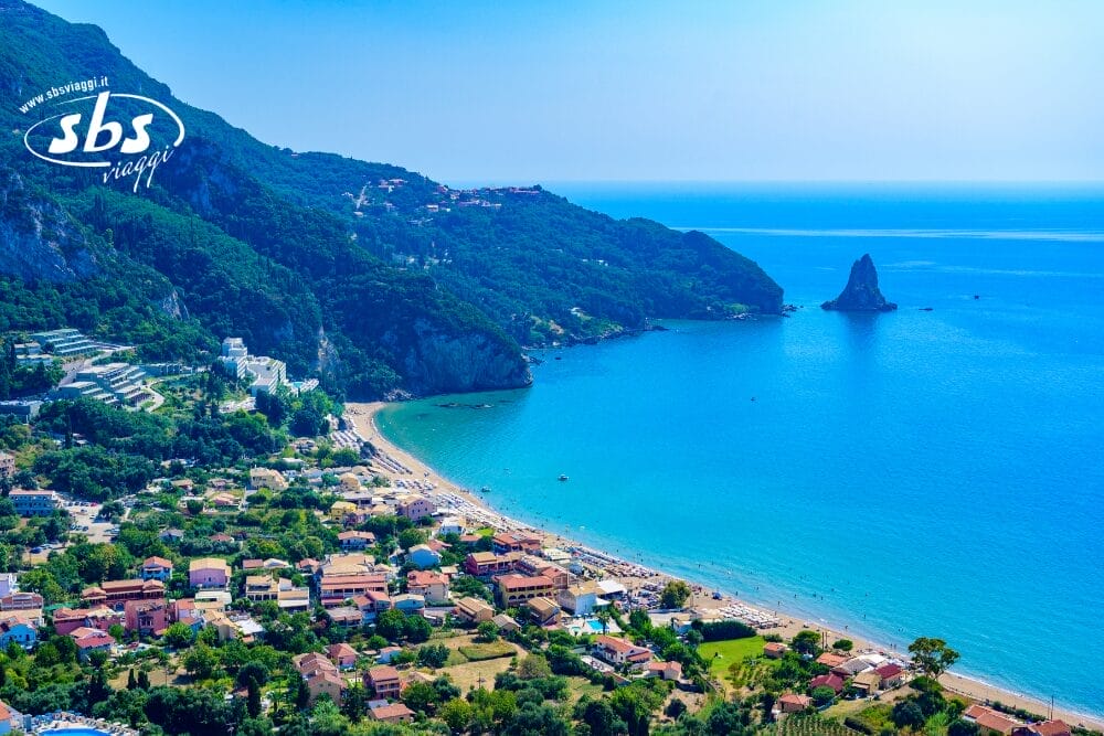 Veduta aerea di un villaggio costiero della Turchia con spiaggia sabbiosa, mare turchese, colline verdeggianti e una grande formazione rocciosa sotto un cielo azzurro e limpido. Il logo "SBS Viaggi" è in alto a sinistra. Volate qui con Turkish Airlines.