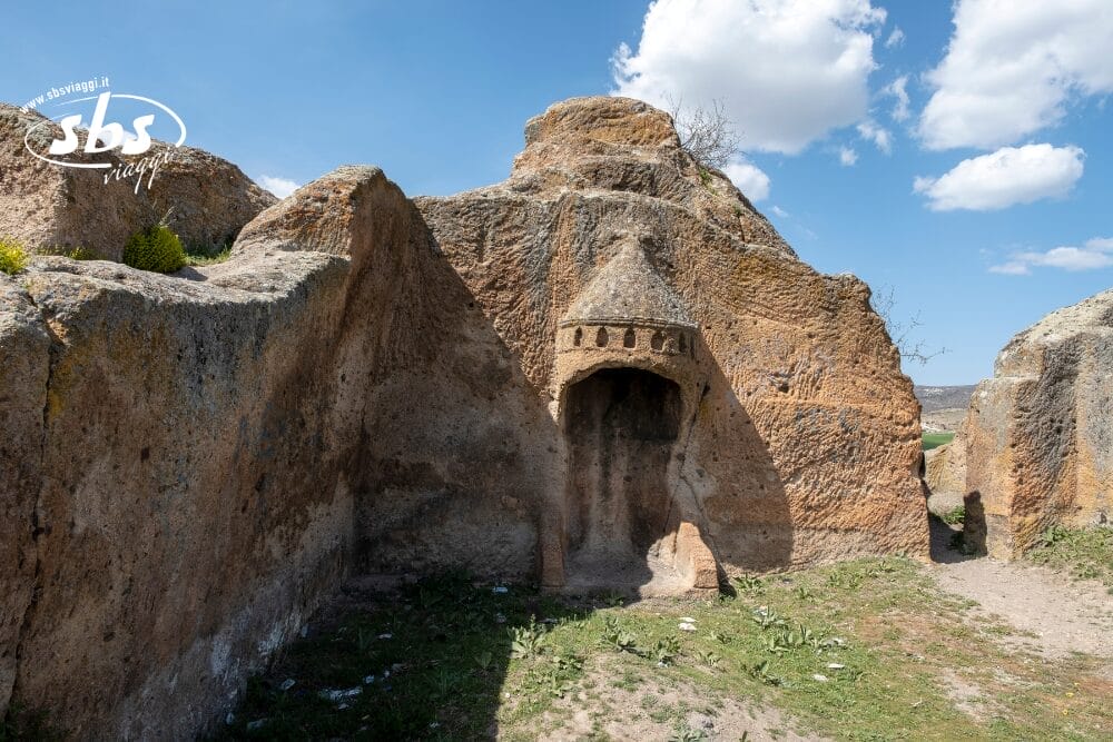 Antica nicchia scavata nella roccia con tetto conico e finestrelle ricavate in una rupe, circondata da un terreno roccioso e aspro sotto un cielo parzialmente nuvoloso: una gemma nascosta in Turchia, da scoprire con SBSViaggi voli inclusi.