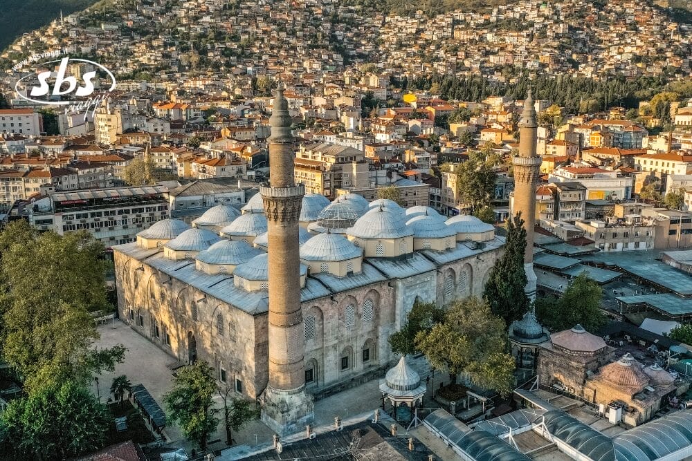 Veduta aerea di una grande moschea storica con cupole multiple e due alti minareti in Turchia, circondata da alberi ed edifici cittadini, case in collina sullo sfondo - scopri con SBSViaggi Stile, voli inclusi.